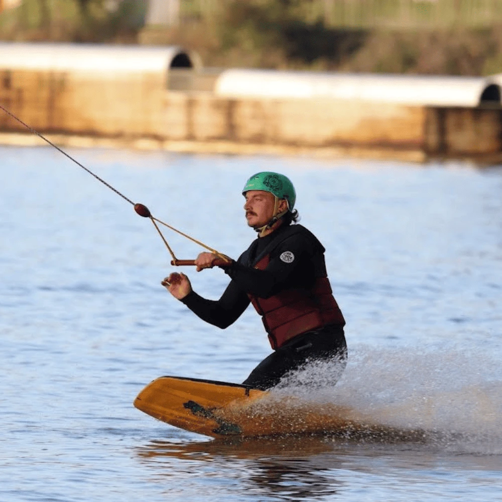 Pratique du kneeboard au wakepark de la Rincerie en Mayenne. Un rider équipé d’un casque vert glisse sur l’eau en plein contrôle avec un câble.