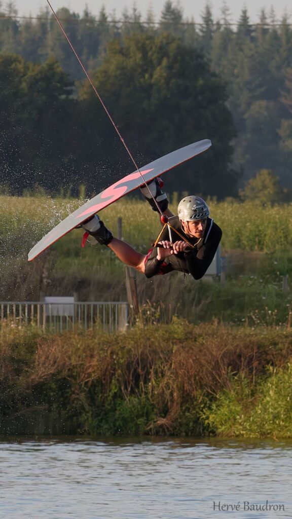 Rider en plein saut acrobatique au Wakepark de la Rincerie, planche inclinée en l'air et casque de protection, lors d'une session de wakeboard.